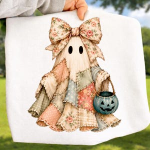 May include: A white fabric item featuring a graphic of a ghost draped in a patchwork quilt made of various floral and plaid patterns in muted pink, blue, and green tones. The ghost wears a large matching floral bow and carries a blue pumpkin bucket.
