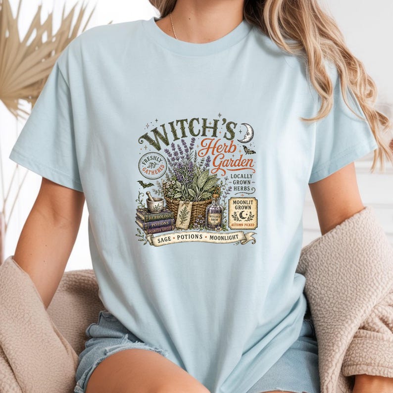May include: A light blue t-shirt featuring a graphic print with the text "Witch's Herb Garden," "Freshly Gathered," "Locally Grown Herbs," "Moonlit Grown," "Autumn Picked," and "Sage, Potions, Moonlight." The design includes herbs, books, and a bottle.