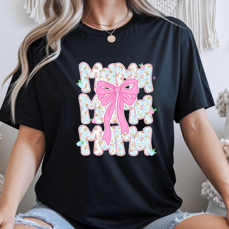 May include: Black t-shirt featuring the word "MAMA" in a floral design, with a pink bow overlay. The floral pattern has a light blue base with small flowers. The bow is positioned centrally over the word "MAMA".