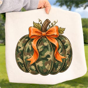 May include: A white fabric item featuring a graphic print of a pumpkin with a camouflage pattern in shades of green, brown, and black. The pumpkin has a brown stem, green leaves, and a large, bright orange bow tied around the top center.