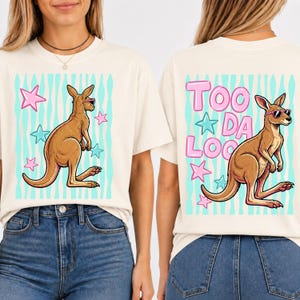 May include: Cream-colored t-shirt featuring a cartoon kangaroo wearing sunglasses, set against a teal striped background with pink and blue stars. The back of the shirt displays the text "TOO DA LOO" with a kangaroo graphic.