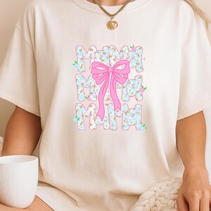 May include: Cream-coloured t-shirt with the word "Mama" in a floral pattern, accented by a pink bow. The design features a light blue floral print on the letters. A gold necklace is visible.