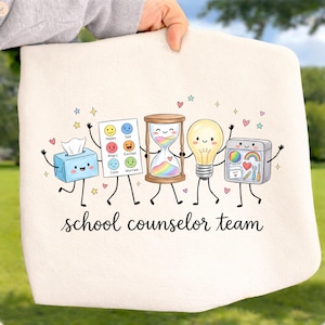 May include: A white tote bag featuring a colorful cartoon illustration of a tissue box, an emotion chart, an hourglass with rainbow sand, a lightbulb, and a supply kit, all with faces and limbs. The text reads school counselor team.
