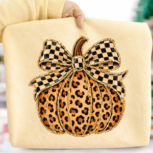 May include: A light yellow sweatshirt featuring a central graphic of a pumpkin with a brown stem and a leopard print pattern. The pumpkin is topped with a large, black and white checkered bow, and the entire design is outlined in gold glitter.