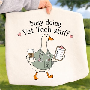 May include: A cream-colored tote bag featuring a cartoon goose dressed in a sage green scrub top with a stethoscope, holding a coffee cup and a clipboard labeled Patient List. The text reads busy doing Vet Tech stuff with two small pink hearts.