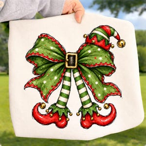 May include: A white fabric item featuring a graphic illustration of a large green bow with red trim and white polka dots. The bow is centered with a black belt buckle and extends into two green and white striped elf legs with red curled shoes.