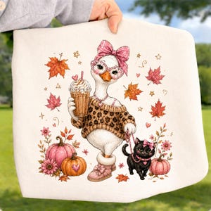 May include: A white fabric item featuring a graphic illustration of a white goose wearing a leopard print sweater, pink boots, a pink bow, and glasses, holding an iced coffee. A black kitten on a pink leash, orange maple leaves, and pumpkins surround it.