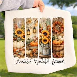May include: A white fabric item featuring four vertical panels with autumn-themed illustrations of sunflowers, pumpkins, lanterns, and wooden textures. The text "Thankful, Grateful, Blessed" is printed in black cursive script at the bottom of the design.