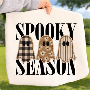 May include: A white fabric item featuring the black text SPOOKY SEASON and three ghost-shaped appliques. The first ghost has a black and white buffalo check pattern, the second has a brown floral print, and the third has brown vertical stripes.