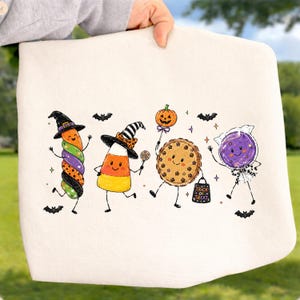 May include: A cream-colored fabric item featuring a colorful Halloween graphic of four anthropomorphic treats: a swirled marshmallow, a candy corn, a pumpkin pie, and a purple lollipop, all wearing hats or holding items with small bats.
