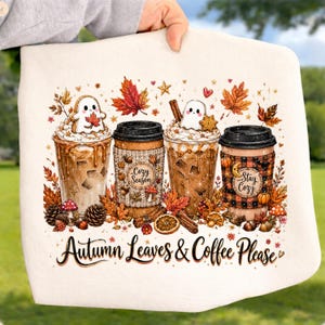 May include: A cream-colored fabric item featuring a graphic of four iced coffee cups decorated with ghosts, autumn leaves, cinnamon sticks, and mushrooms. The text "Autumn Leaves & Coffee Please" is printed in a dark script font at the bottom.