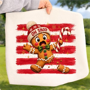May include: A white fabric item featuring a gingerbread character wearing a red and white knit hat with the text OH SNAP. The character has a green bow tie, holds a red and white candy cane, and has a broken leg. Red brush strokes and gold snowflakes decorate the background.