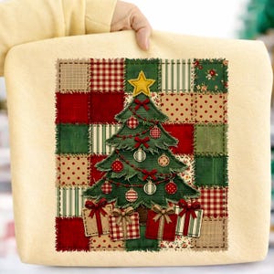 May include: A cream-colored sweatshirt featuring a central patchwork-style graphic of a green Christmas tree decorated with red and white ornaments, bows, and a yellow star, surrounded by a grid of red, green, and beige patterned fabric squares.