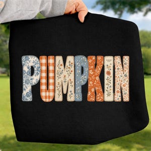 May include: A black sweatshirt featuring the word PUMPKIN in large block letters. Each letter has a unique pattern, including blue floral, orange gingham, wildflower prints, and solid orange, all outlined with a decorative stitched border.