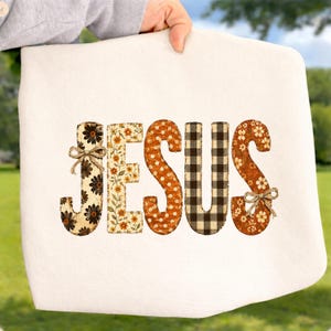 May include: A white fabric item featuring the word JESUS in large, decorative letters. Each letter has a unique pattern: floral prints, orange polka dots, and a black and white buffalo check. Two small jute twine bows are attached to the letters.