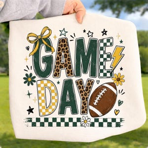 May include: A white sweatshirt featuring the text GAME DAY in a mix of dark green, leopard print, and yellow checkered patterns. The design includes a yellow bow, a lightning bolt, a brown football, and small flower and star accents.
