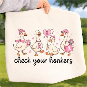 May include: A cream-colored fabric item featuring a graphic of four cartoon geese with pink breast cancer awareness ribbons. The geese wear hats, scarves, and hold a coffee cup, a heart balloon, and a tote bag. The text reads, check your honkers.