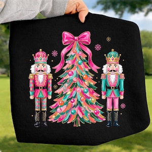 May include: A black sweatshirt featuring a graphic print of a stylized Christmas tree decorated with pink, teal, and gold ornaments, topped with a large pink bow, flanked by two nutcracker figures in matching pink and teal uniforms with crowns.