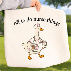 May include: A cream-colored tote bag featuring a graphic of a white goose dressed as a nurse, wearing a cap, stethoscope, and scrubs, holding a coffee cup and clipboard. The text above the goose reads, off to do nurse things.