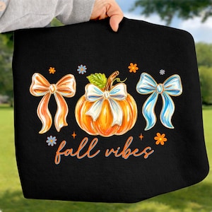 May include: A black sweatshirt featuring a central orange pumpkin with a white bow, flanked by an orange bow on the left and a blue bow on the right. Small orange and blue flowers surround the design, with the text fall vibes in orange script.