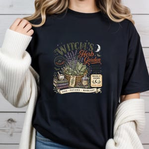 May include: A black t-shirt featuring a graphic print with the text Witch's Herb Garden, Moonlit Grown, Autumn Picked, Sage, Potions, and Moonlight. The design includes a basket of herbs, a mortar and pestle, stacked books, and a glass bottle.