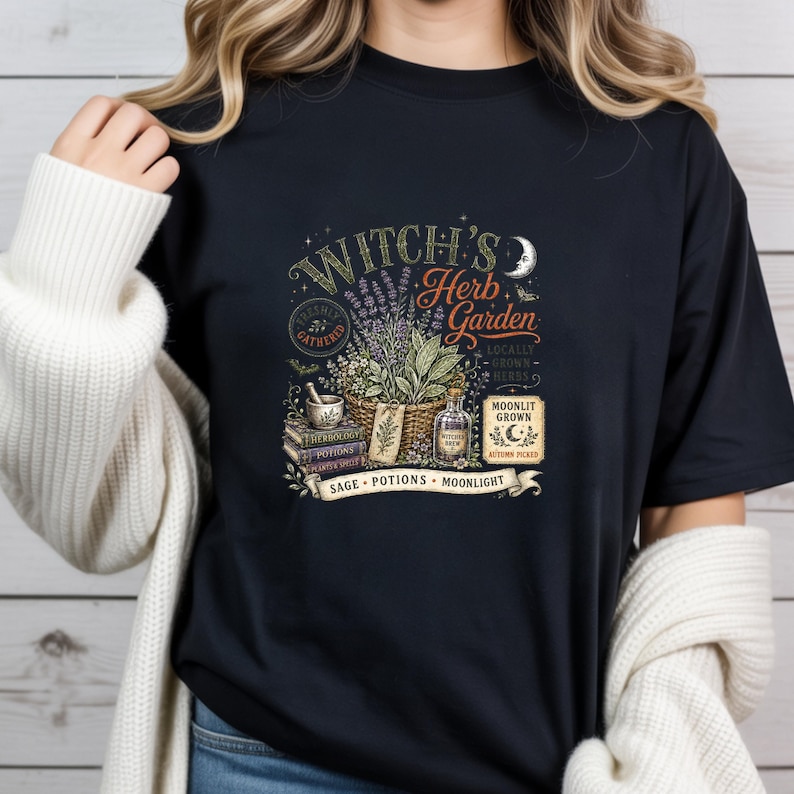 May include: A black t-shirt featuring a graphic print with the text Witch's Herb Garden, Moonlit Grown, Autumn Picked, Sage, Potions, and Moonlight. The design includes a basket of herbs, a mortar and pestle, stacked books, and a glass bottle.