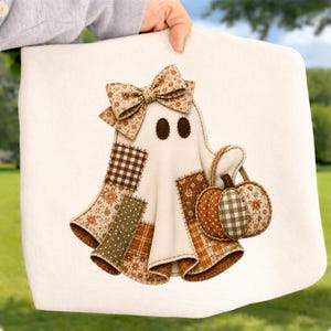 May include: A white fabric square featuring a ghost applique with a floral bow. The ghost is decorated with brown and green patches in gingham, polka dot, and floral patterns. It holds a small pumpkin with matching fabric textures and colors.