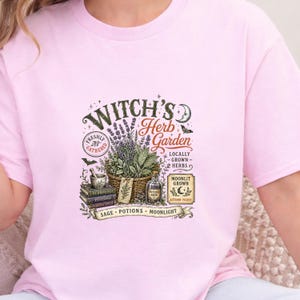 May include: A light pink t-shirt featuring a graphic print with the text Witch's Herb Garden. The design includes a basket of herbs, a mortar and pestle, stacked books labeled Herbology, Potions, and Plants & Spells, and a bottle of potion.