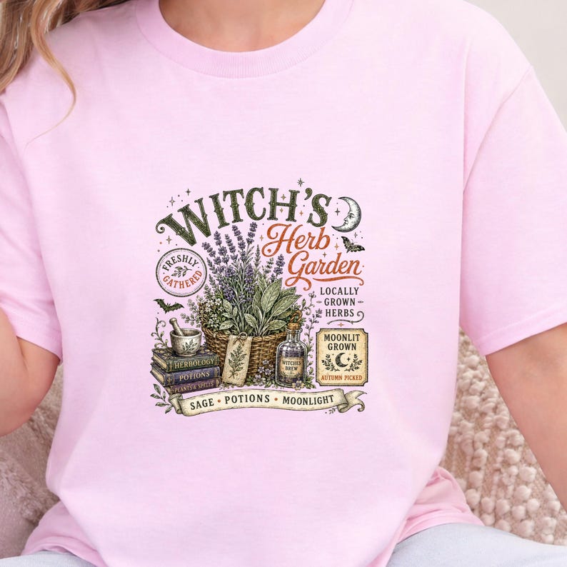 May include: A light pink t-shirt featuring a graphic print with the text Witch's Herb Garden. The design includes a basket of herbs, a mortar and pestle, stacked books labeled Herbology, Potions, and Plants & Spells, and a bottle of potion.