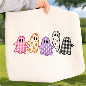 May include: A white fabric item featuring a row of five ghost-shaped appliques. The ghosts have distinct patterns: pink gingham, orange pumpkins, purple polka dots, white with black stars, and black and white buffalo plaid, all with black eyes.