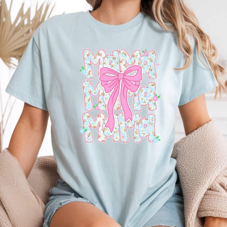 May include: Pale blue t-shirt featuring the word "Mama" in a floral design, repeated three times. A large pink bow is centered over the text. The shirt is a casual style.