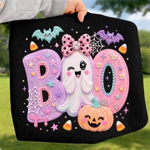 May include: A black sweatshirt featuring a colorful graphic with the text BOO. The design includes a winking ghost with a pink polka dot bow, a smiling orange pumpkin, two bats, spiderwebs, stars, hearts, and candy corn accents.