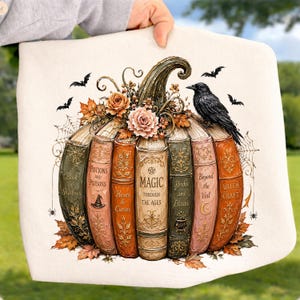 May include: A cream-colored fabric item featuring a graphic of a pumpkin constructed from stacked vintage books. The books are titled Book of Shadows, Potions and Poisons, Hexes & Curses, Magic Through the Ages, Herbs and Elixirs, Beyond the Veil, and Witch Craft. A black crow, bats, and spiderwebs surround the pumpkin.