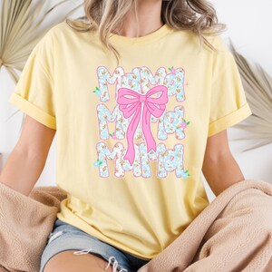 May include: A pale yellow t-shirt featuring the word "Mama" in a floral design, repeated three times vertically. A large pink bow is centered over the word. The shirt has short sleeves and a loose fit.