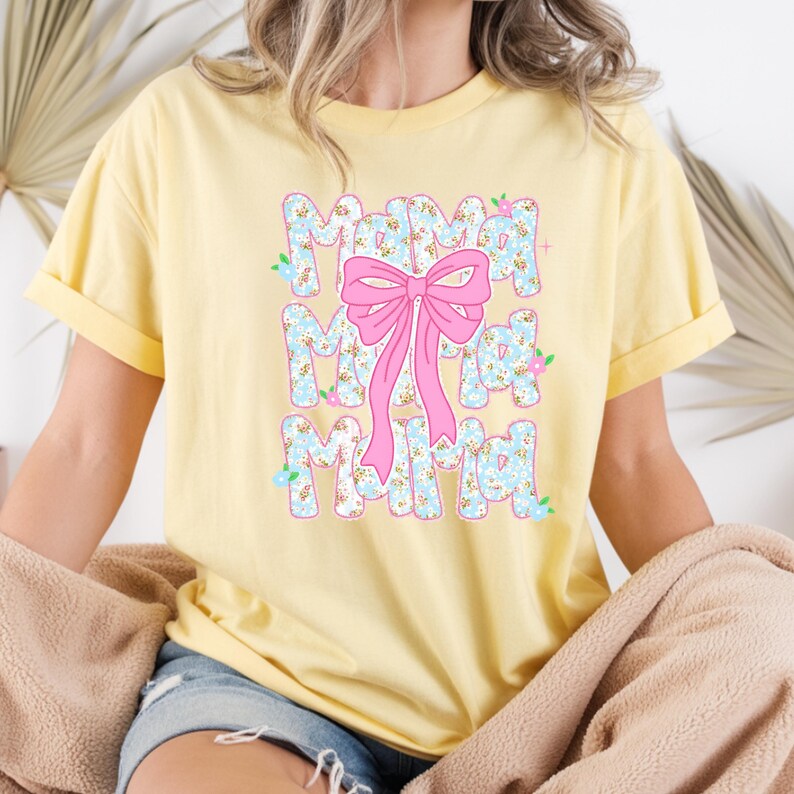 May include: A pale yellow t-shirt featuring the word "Mama" in a floral design, repeated three times vertically. A large pink bow is centered over the word. The shirt has short sleeves and a loose fit.