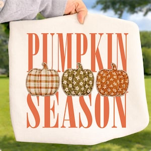 May include: A white fabric item featuring the orange text PUMPKIN SEASON in a tall font. Between the words are three pumpkin illustrations: one with an orange and white plaid pattern, one green with a white floral print, and one orange with a white floral print.