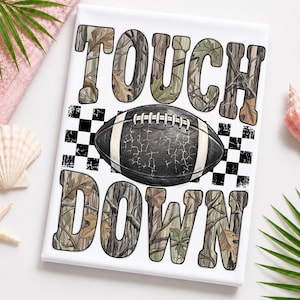 May include: A rectangular white canvas print featuring the text TOUCH DOWN in large block letters filled with a camouflage pattern of brown and green leaves. A black football with white stripes and a cracked texture sits between the words.