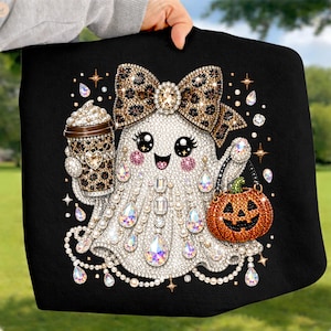 May include: A black t-shirt featuring a graphic of a white ghost adorned with sparkling rhinestones. The ghost wears a large leopard print bow, holds a leopard print coffee cup, and carries an orange jack-o'-lantern bucket with a green stem.