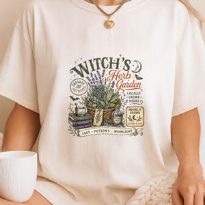 May include: A cream-colored t-shirt featuring a graphic print with the text "Witch's Herb Garden". The design includes a basket of herbs, a mortar and pestle, stacked books, a glass bottle, and small illustrations of a moon, stars, and bats.