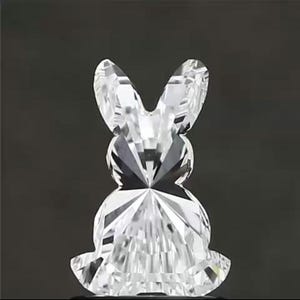 May include: A clear, faceted crystal bunny figurine. The bunny is depicted in a sitting position with long ears and a round body. The crystal reflects light, creating a sparkling effect. The background is a dark gray.