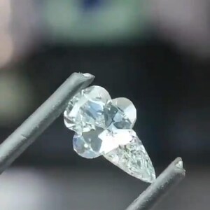 May include: A clear, faceted diamond shaped like an ice cream cone, held by silver tweezers. The diamond has a cone and three scoops, reflecting light. The background is blurred, focusing attention on the gemstone.