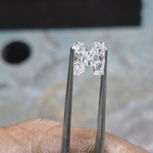 May include: Two clear, rectangular-cut diamonds are held by metal tweezers. The diamonds are positioned close together, showcasing their facets and brilliance. The background is blurred, focusing attention on the gemstones.