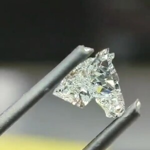 May include: A small, irregularly shaped, clear gemstone is held by a pair of silver tweezers. The gemstone has many facets, reflecting light. The background is blurred, with hints of yellow and blue.