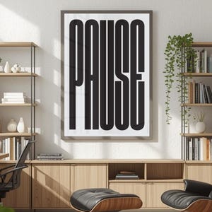May include: A rectangular wall art print featuring the word PAUSE in bold, elongated black sans-serif capital letters centered on a white background, enclosed within a thin dark frame, displayed on a wall above a wooden cabinet.