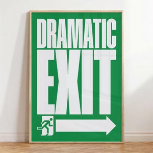 May include: A framed poster with a green background and white text that reads "DRAMATIC EXIT." The poster includes an exit sign graphic with a person running towards an arrow. The frame is a light wood color.