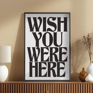 Puede incluir: Impresión artística enmarcada con la frase "WISH YOU WERE HERE" en letras negras sobre fondo blanco. La obra se exhibe sobre un mueble de madera, con una lámpara y jarrones decorativos.