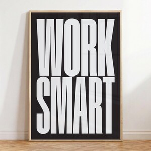 Op de afbeelding: Een rechthoekige wandkunstprint met een zwarte achtergrond en de woorden WORK SMART in grote, vette, witte schreefloze hoofdletters. De print is ingelijst in een dunne, lichtgekleurde houten lijst met een strakke en eenvoudige uitstraling.