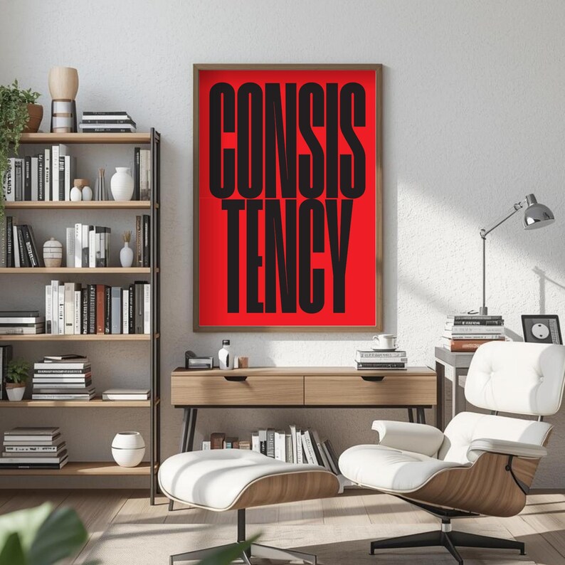 Op de afbeelding: Een ingelijste kunstprint met het woord "CONSISTENCY" in grote, vette, zwarte letters op een helderrode achtergrond. De print wordt getoond in een moderne woonkamer met een houten boekenplank, bureau en een witte loungestoel.