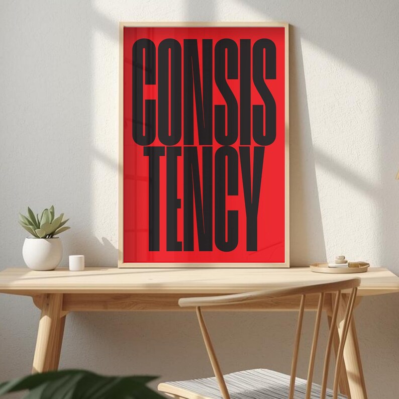 Op de afbeelding: Een ingelijste print met het woord "CONSISTENCY" in grote, zwarte hoofdletters op een felrode achtergrond. De print zit in een licht houten frame en staat op een houten bureau.