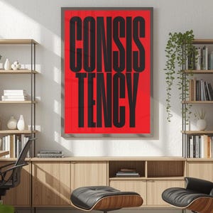 Op de afbeelding: Een ingelijste poster met het woord "CONSISTENCY" in grote, vette, zwarte letters tegen een felrode achtergrond. De poster wordt weergegeven op een muur boven een houten kast met planken en een zwarte stoel.